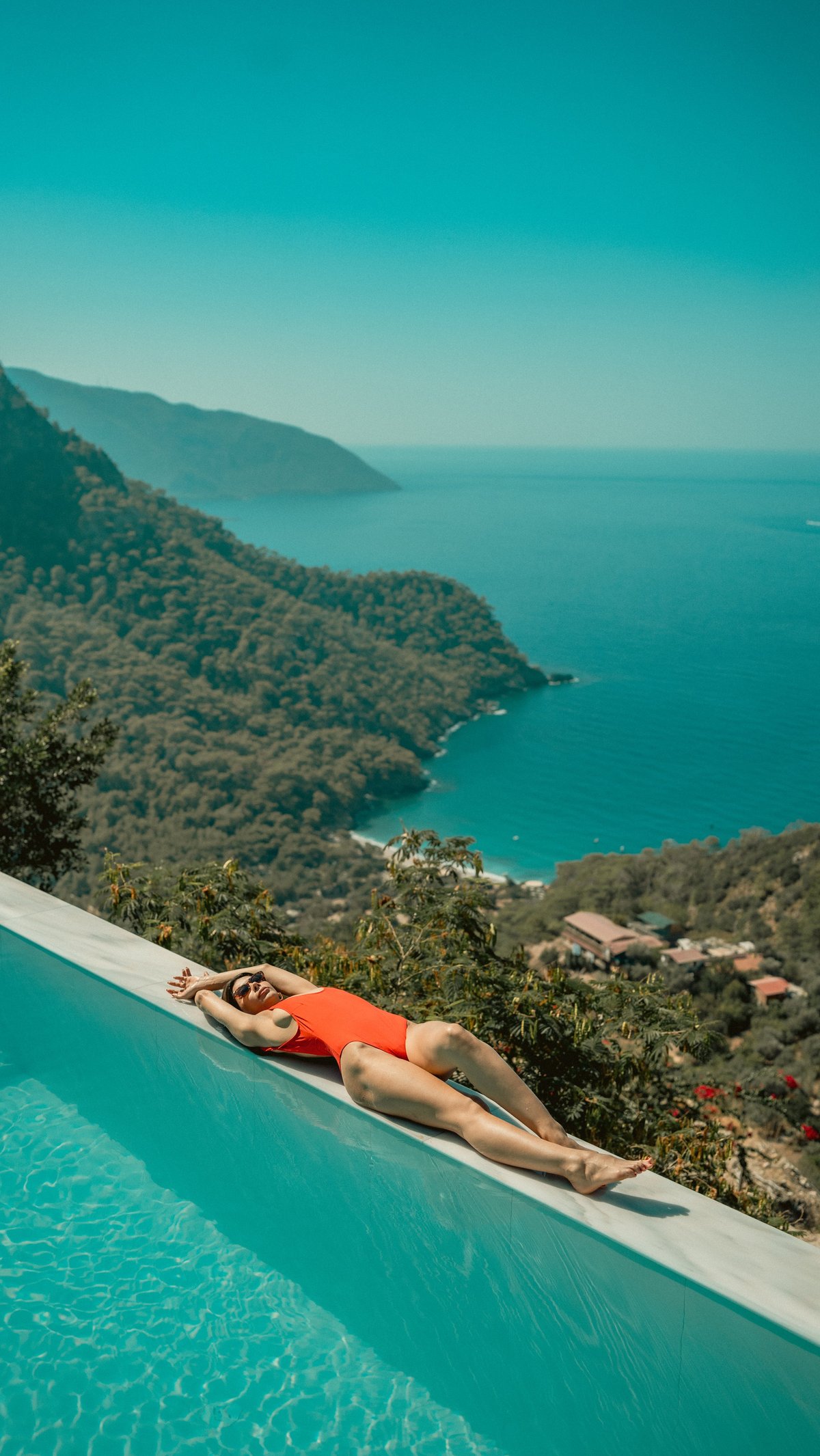A woman sunbathes by a luxurious infinity pool overlooking a breathtaking coastal landscape with lush green mountains and clear blue sea